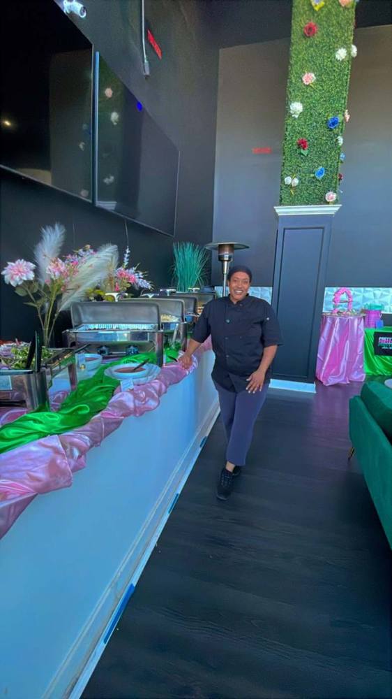 Chef-owner Charayle smiling in her black chef coat beside a pink-and-green catered buffet